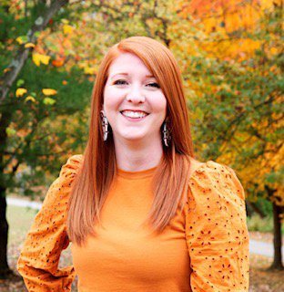 A young woman with long red hair smiling outdoors during autumn, wearing an orange top with puffed sleeves and dangling earrings, standing in front of colorful fall trees.