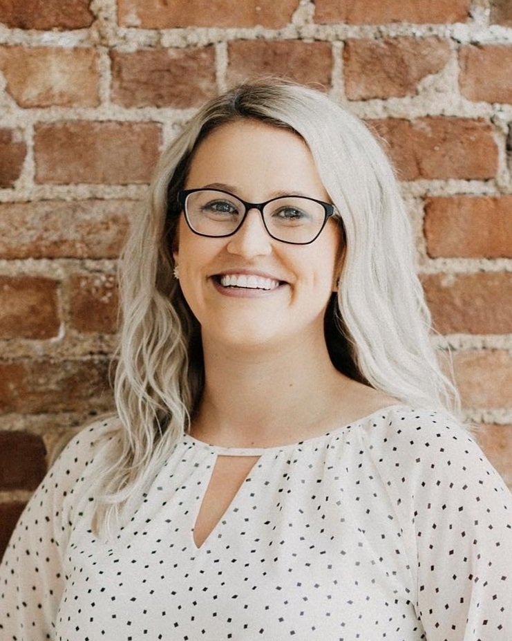 A woman with blonde hair wearing glasses and a white polka dot blouse, standing in front of a brick wall and smiling.