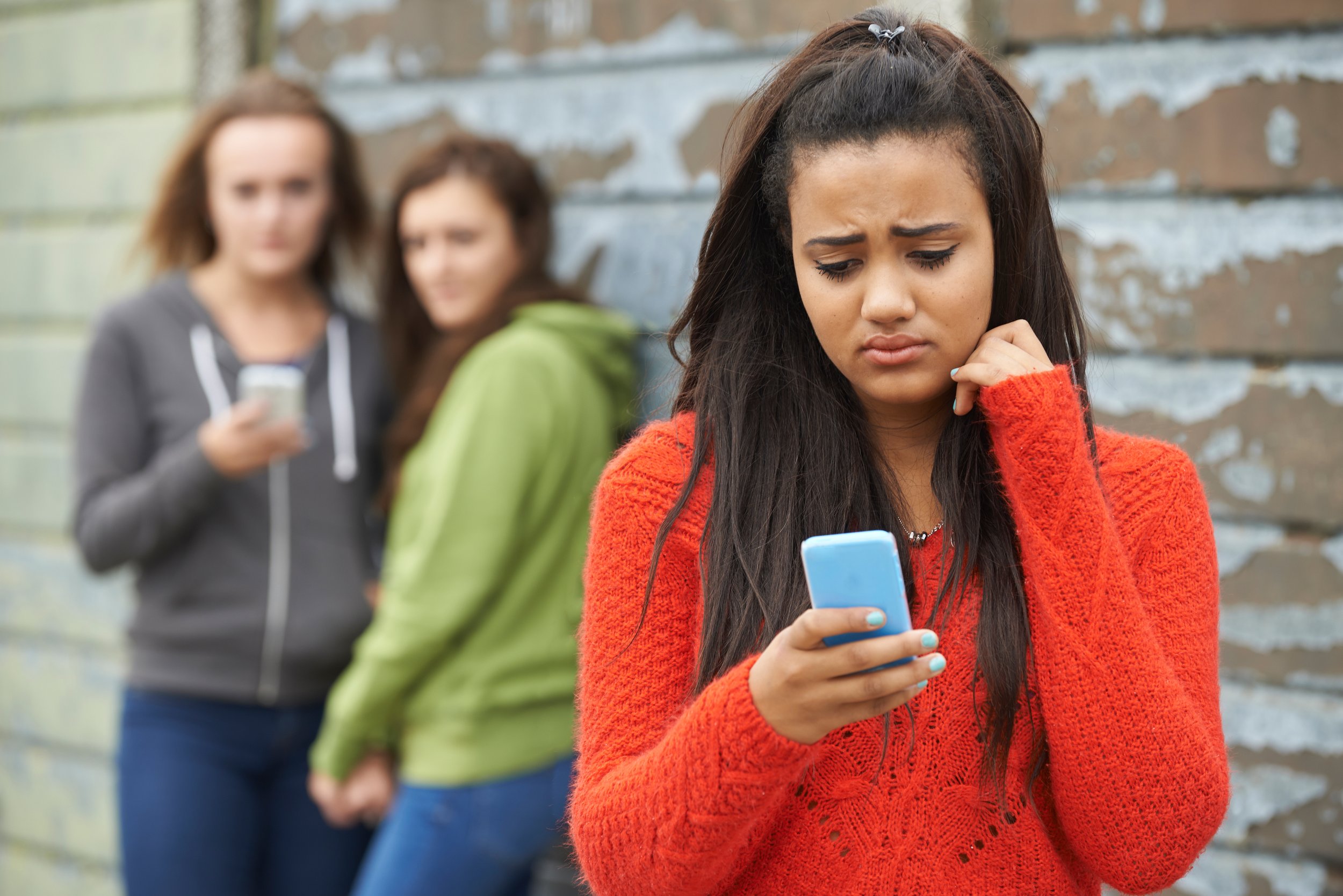 Girl in red sweater looking upset at her phone while two girls in the background are also using their phones, standing against a wall.